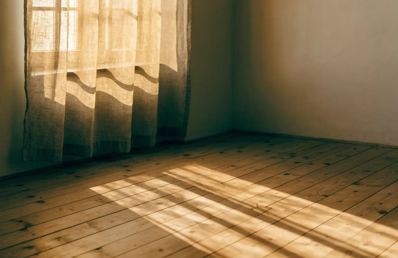 Low golden sunrise light streaming through a tall window onto a wide wooden floor, a sheer linen curtain glowing amber beside the window.